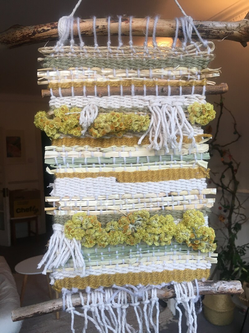 K&ouml;nnte beinhalten: Ein gewebter Wandbehang mit Naturfasern und getrockneten Blumen. Der Behang ist aus verschiedenen Texturen und Farben gefertigt, darunter Wei&szlig;, Beige, Gr&uuml;n und Gelb. Der Behang ist an zwei Holzst&ouml;cken befestigt.