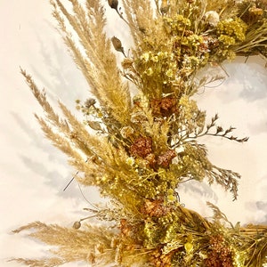 May include: A dried floral wreath with a variety of dried flowers and grasses in shades of beige, brown, and white. The wreath is made with a natural base and is perfect for adding a touch of rustic charm to any space.