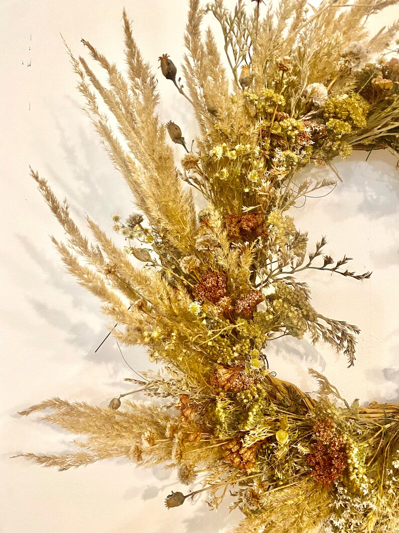 May include: A dried floral wreath with a variety of dried flowers and grasses in shades of beige, brown, and white. The wreath is made with a natural base and is perfect for adding a touch of rustic charm to any space.