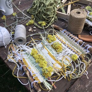 K&ouml;nnte beinhalten: Nahaufnahme einer gewebten Wandbeh&auml;nge aus nat&uuml;rlichen Materialien. Die Beh&auml;nge hat ein wei&szlig;-gelbes Streifenmuster mit getrockneten gelben Blumen, die in das Design eingewebt sind. Die Beh&auml;nge ist von nat&uuml;rlichem Garn und St&ouml;cken umgeben.