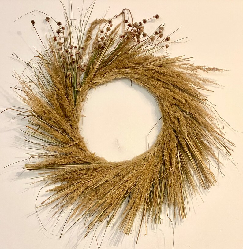 May include: A natural-toned wreath made of dried grasses and flowers. The wreath is circular and has a rustic, bohemian style.