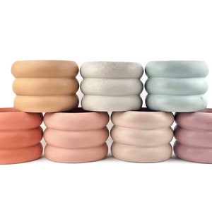 May include: Set of six concrete planters in various pastel colors, each with a unique rippled design. The planters are arranged in a row, with the largest planter in the back and the smallest planter in the front.