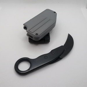 May include: A black karambit knife with a curved blade and finger ring, paired with a gray sheath. The knife has a black handle and is designed for tactical use. The sheath is gray and has a rectangular shape.