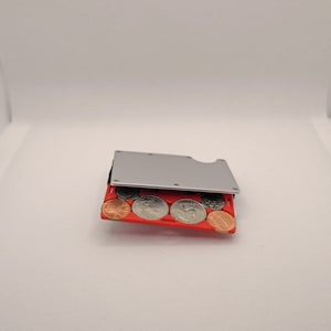 May include: A silver metal wallet with a red coin holder, filled with various coins. The wallet has a sleek, minimalist design and a small cutout on one side. The coins are a mix of pennies and quarters.