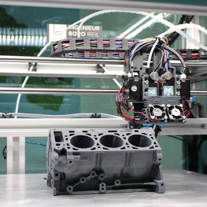 May include: A 3D printer in operation, with a gray engine block model in progress. The printer head, with multiple wires and fans, is positioned above the model. The machine is set up on a metal surface.