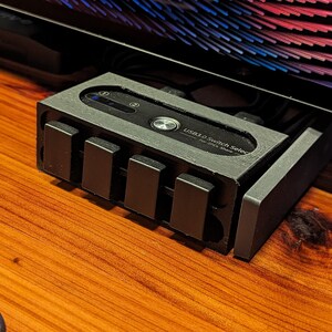 May include: A black USB 3.0 switch selector with four rectangular buttons and a small LED indicator. The device is labeled "USB 3.0 Switch Selector For 2PC Share". It sits on a wooden surface, partially beneath a monitor.