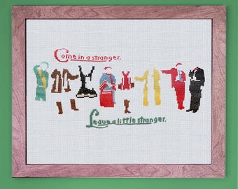 May include: Cross-stitch embroidery in a wooden frame with the text "Come in a stranger, leave a little stranger."  The embroidery depicts a group of people in various colorful outfits.