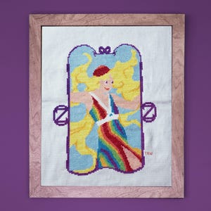 May include: Cross-stitch artwork of a blonde figure in a rainbow dress, framed in light wood. The figure is inside a blue arch with yellow accents and the letter "Z" on each side. The artwork is on a white background.