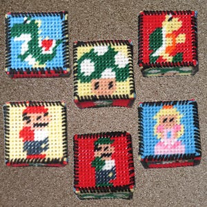 May include: Six small, square, embroidered pillow-like boxes with different characters from the Super Mario Bros. video game. The characters include Mario, Luigi, Princess Peach, a green mushroom, a turtle, and a dragon.