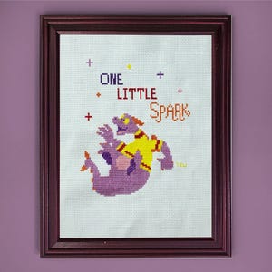 May include: Cross-stitch art in a dark wood frame. The artwork features a purple dragon-like character wearing a yellow shirt. Text reads "ONE LITTLE SPARK" in blue, red, and orange. Plus signs and a yellow star are also included.