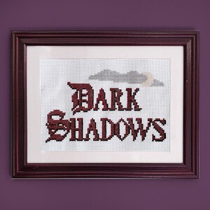 May include: A framed cross-stitch with the words "DARK SHADOWS" in red lettering. The cross-stitch is on white fabric and is framed in a dark wood frame.
