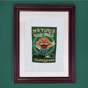 Op de afbeelding: Een ingelijst kruissteek met een cartoon kikker met de tekst "M.A.T.U.A.D'S WILD JUICE" en "Fantastico!"