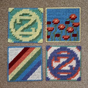 May include: Four square, handmade needlepoint coasters. Designs include a stylized letter "Z" in yellow and green, red flowers on blue, diagonal stripes in various colors, and a blue and white design with orange accents. Each coaster has a contrasting border.