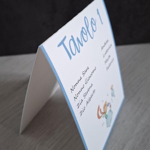 Può includere: Un segnaposto bianco con bordi e testo azzurro. Il segnaposto è piegato e mostra le parole "Tavolo 1" in corsivo azzurro. I nomi sono elencati sul segnaposto, insieme a una piccola illustrazione.