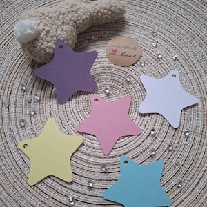 Può includere: Cinque etichette regalo a forma di stella in diversi colori: viola, rosa, giallo, blu e bianco. Le etichette hanno un foro perforato nella parte superiore e sono su uno sfondo intrecciato. Una piccola etichetta marrone con il testo "fatto a mano con amore" è visibile sullo sfondo.