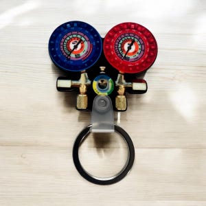 May include: A red and blue gauge keychain with a silver ring. The gauges are labeled with numbers and have a black background. The keychain is shaped like a set of valves.