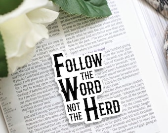 以下が含まれることがあります： 太字のスタイリッシュなセリフ体で「FOLLOW THE WORD NOT THE HERD」という黒い文字が書かれた白いダイカットステッカー。印刷されたテキストが見える本の開いたページの上に、白い花と一緒に配置されています。