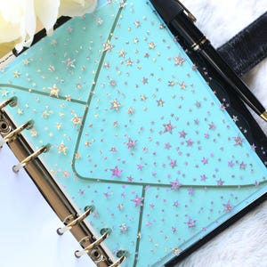 May include: A planner with a light blue cover featuring a star pattern. The planner is open, revealing a gold-colored binder mechanism and white pages. A black pen rests on the planner, and a white flower is in the background.