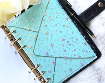 May include: A planner with a light blue cover featuring a star pattern. The planner is open, revealing a gold-colored binder mechanism and white pages. A black pen rests on the planner, and a white flower is in the background.
