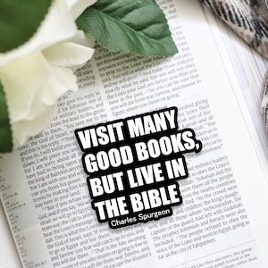 May include: A black and white sticker with the text "Visit many good books, but live in the Bible" and the name "Charles Spurgeon" below the text. The sticker is placed on an open book with white pages.