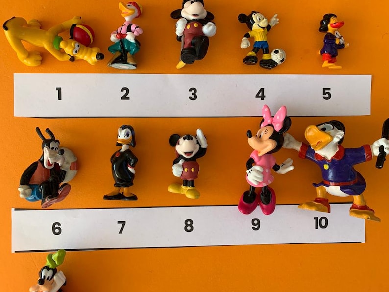 K&ouml;nnte beinhalten: Zehn kleine Plastikfiguren von Disney-Charakteren, darunter Mickey Mouse, Minnie Mouse, Donald Duck, Goofy und Pluto. Die Figuren sind von 1 bis 10 nummeriert.
