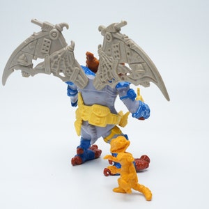 May include: A gray and blue action figure with wings and a yellow belt stands on a white surface. A smaller orange action figure with a tail stands in front of the larger figure.