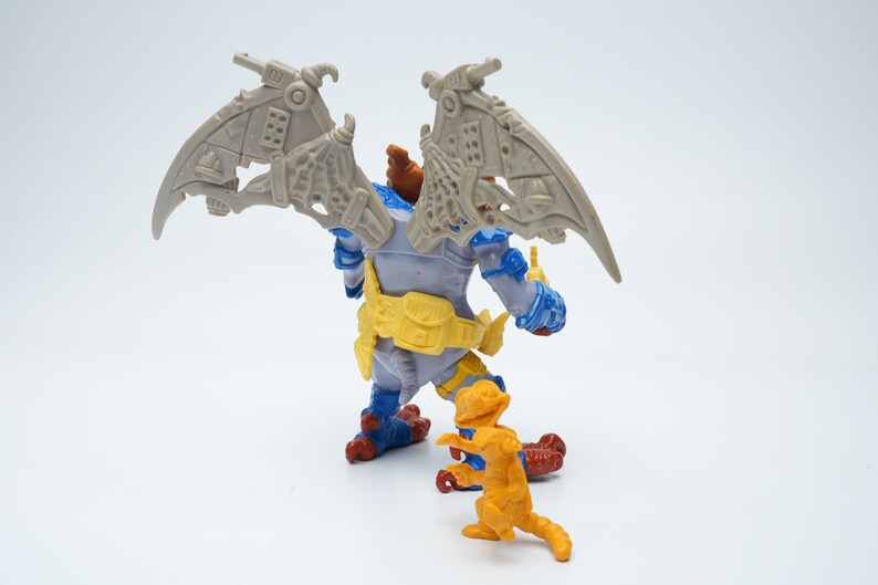 May include: A gray and blue action figure with wings and a yellow belt stands on a white surface. A smaller orange action figure with a tail stands in front of the larger figure.