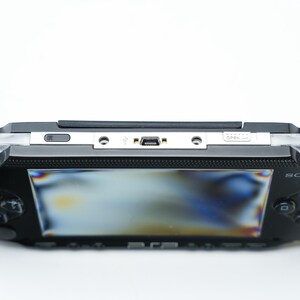 May include: Black Sony PSP handheld game console. The device has a screen, control buttons, and a directional pad. The top edge features a silver panel with ports and a power button. The Sony logo is visible on the device.
