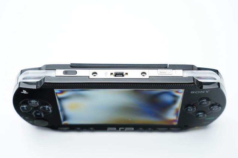 May include: Black Sony PSP handheld game console. The device has a screen, control buttons, and a directional pad. The top edge features a silver panel with ports and a power button. The Sony logo is visible on the device.