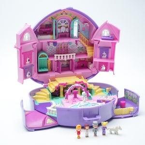 Può includere: Una casa delle bambole vintage, aperta, rosa e viola con più stanze, scale e una fontana. La casa delle bambole ha la forma di un cuore e include figurine in miniatura. Il tema generale è un giocattolo classico.