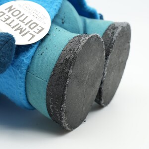 Puede incluir: Primer plano de un juguete de peluche azul con pies de tela negra. El juguete tiene una etiqueta que dice "EDICI&Oacute;N LIMITADA" con el texto "COLLECTA THE ORIGINAL" y "AVAILABLE NOW".