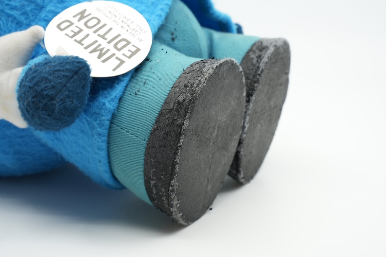 Puede incluir: Primer plano de un juguete de peluche azul con pies de tela negra. El juguete tiene una etiqueta que dice "EDICI&Oacute;N LIMITADA" con el texto "COLLECTA THE ORIGINAL" y "AVAILABLE NOW".