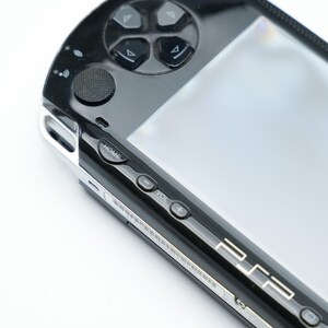 May include: Close-up of a black handheld gaming console with a silver trim. The console features a directional pad, buttons, and a screen. The word "HOME" is visible, along with volume controls and the PSP logo.