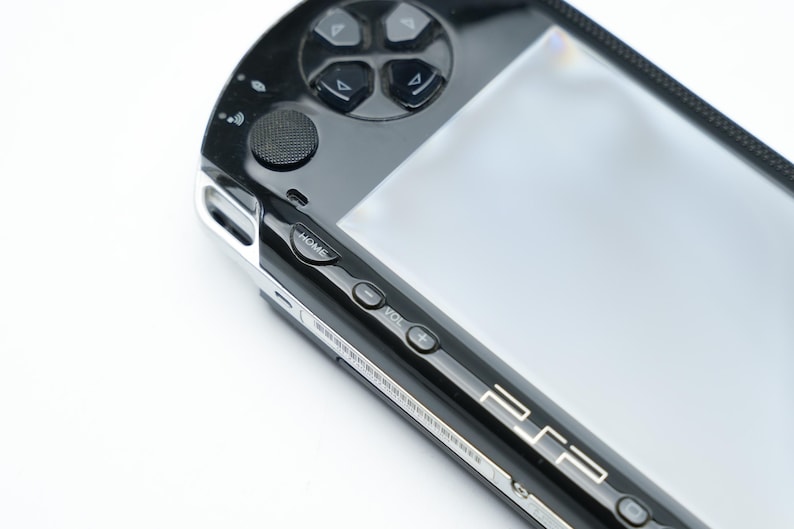May include: Close-up of a black handheld gaming console with a silver trim. The console features a directional pad, buttons, and a screen. The word "HOME" is visible, along with volume controls and the PSP logo.