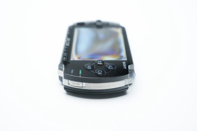 May include: Black Sony PSP handheld game console. The device has a screen, directional buttons, and action buttons. The console has a silver trim and the Sony logo is visible.