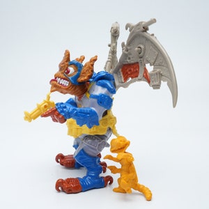 May include: A blue and brown action figure with a yellow gun and a gray wing. The figure is standing next to a smaller orange action figure.