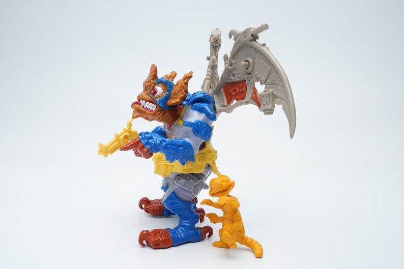 May include: A blue and brown action figure with a yellow gun and a gray wing. The figure is standing next to a smaller orange action figure.