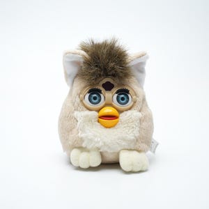 May include: A tan, brown, and white Furby toy. It features large blue eyes, a yellow beak, and a brown tuft of fur. This plush toy is a popular collectible item.
