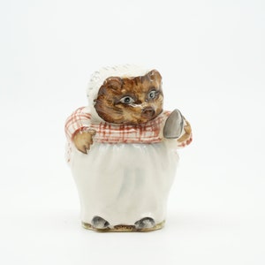 Puede incluir: Una figurita de cerámica de un erizo marrón y blanco que lleva un vestido a cuadros rojo y blanco y sostiene una pequeña plancha. La figurita está de pie sobre un fondo blanco.
