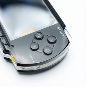 May include: A close-up of a black Sony PSP handheld gaming console. The device features a glossy black finish, silver trim, and a screen. Visible are the directional buttons, action buttons, and the Sony logo. The power button is illuminated in green.