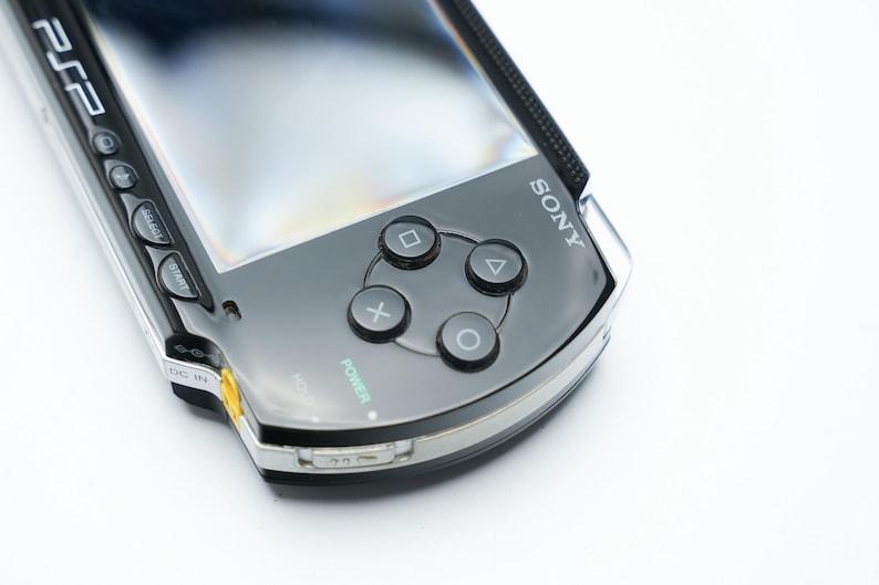 May include: A close-up of a black Sony PSP handheld gaming console. The device features a glossy black finish, silver trim, and a screen. Visible are the directional buttons, action buttons, and the Sony logo. The power button is illuminated in green.