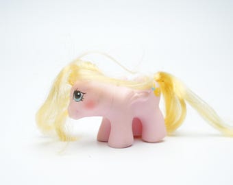 Puede incluir: Un juguete de pony rosa con una melena y cola amarillas. El pony tiene ojos azules y una nariz rosa.