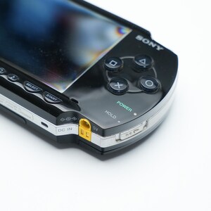 May include: A black Sony PSP handheld gaming console. The device features a glossy black finish, a screen, and control buttons including a directional pad, action buttons, and start/select buttons. The power button is labeled and illuminated.