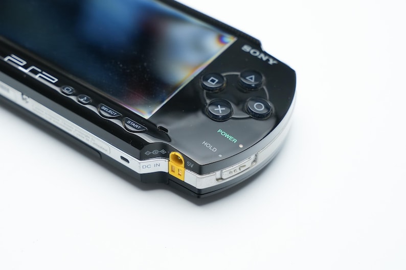 May include: A black Sony PSP handheld gaming console. The device features a glossy black finish, a screen, and control buttons including a directional pad, action buttons, and start/select buttons. The power button is labeled and illuminated.