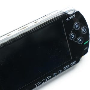 May include: A black Sony PSP handheld gaming console. The device features a black screen, directional buttons, and action buttons. The Sony logo is visible on the top, and the PSP logo is on the bottom. The console is set against a white background.