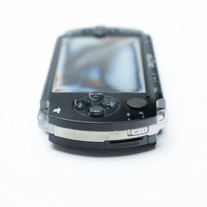 May include: A black handheld gaming console with a silver trim. The device features a screen, directional pad, action buttons, and a small analog stick. The console is on a white background.