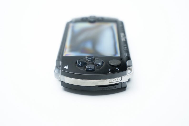 May include: A black handheld gaming console with a silver trim. The device features a screen, directional pad, action buttons, and a small analog stick. The console is on a white background.