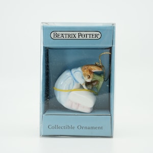 Puede incluir: Un adorno coleccionable de Beatrix Potter con Peter Rabbit con un vestido azul sosteniendo una zanahoria. El adorno está empaquetado en una caja azul con el texto "Beatrix Potter" y "Adorno coleccionable".