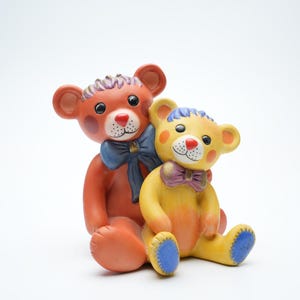 Puede incluir: Dos osos de peluche de cerámica, uno naranja y otro amarillo, con detalles azules y morados. El oso naranja lleva un lazo azul y el oso amarillo lleva un lazo morado.
