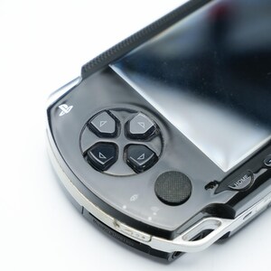 May include: A close-up of a black and silver handheld gaming console. The console features a directional pad with four buttons, a circular button, and a screen. The buttons are black, and the console has a silver trim.